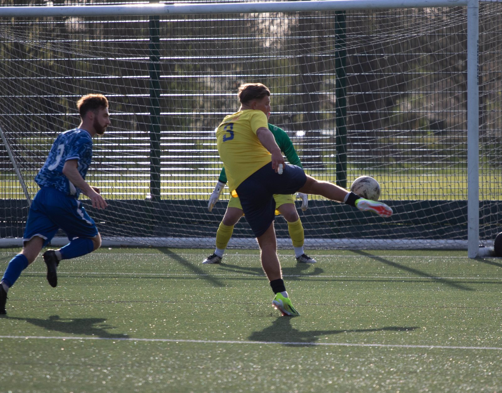 Football attacker shoots towards goal as defenders close in.